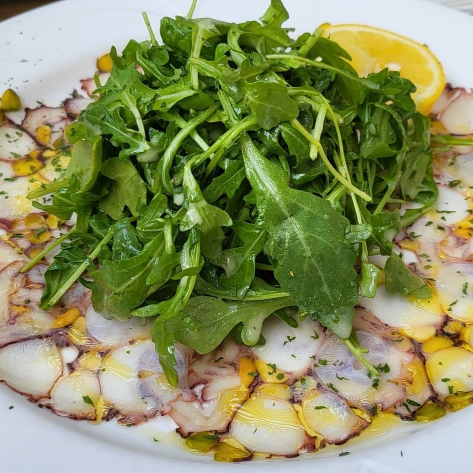 Thinly sliced octopus carpaccio with arugula, olive oil, and a lemon wedge on a white plate.