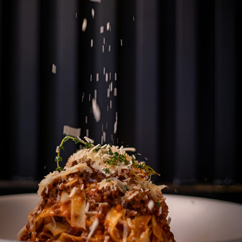 Parmesan cheese being sprinkled over a plate of fresh Italian pasta with meat sauce and herbs.