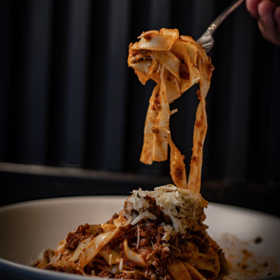 Fork lifting tagliatelle pasta with bolognese sauce and grated cheese from a white bowl.